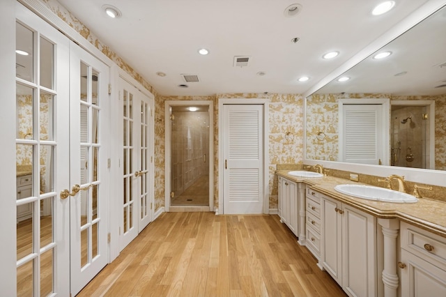 bathroom with french doors, a shower stall, wood finished floors, wallpapered walls, and double vanity