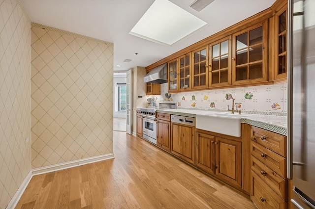 kitchen featuring appliances with stainless steel finishes, a sink, wall chimney range hood, brown cabinets, and light wood-type flooring