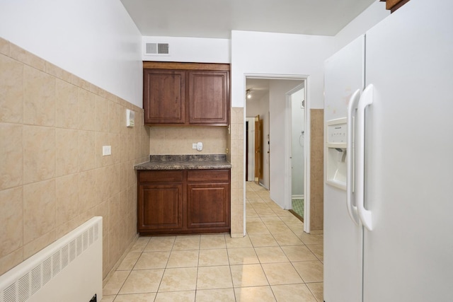 kitchen featuring white fridge with ice dispenser, radiator, light tile patterned floors, dark countertops, and tile walls