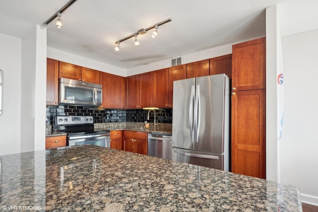 kitchen featuring stainless steel appliances, decorative backsplash, dark stone countertops, a sink, and track lighting