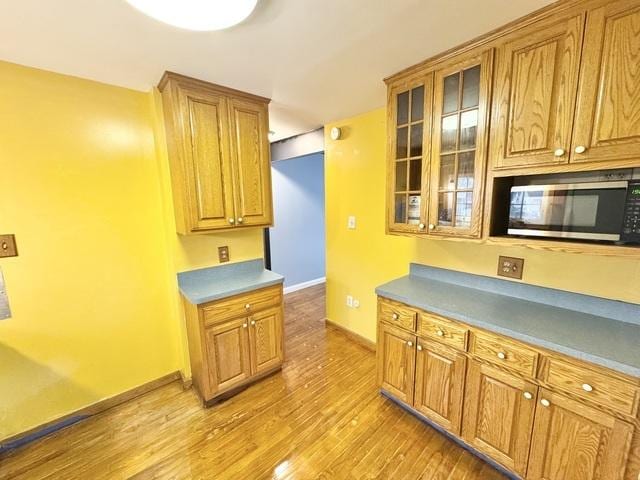 kitchen featuring stainless steel microwave, light wood finished floors, baseboards, glass insert cabinets, and brown cabinets