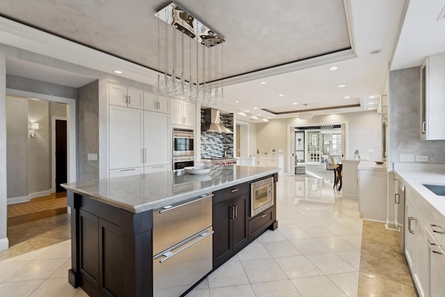 kitchen featuring wall chimney exhaust hood, a raised ceiling, appliances with stainless steel finishes, light tile patterned flooring, and light stone countertops