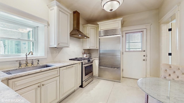 kitchen with appliances with stainless steel finishes, wall chimney range hood, a sink, light stone counters, and light tile patterned floors