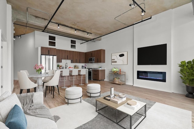 living room featuring rail lighting, a towering ceiling, light wood-style flooring, and a glass covered fireplace