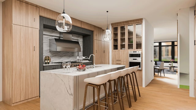 kitchen featuring wall chimney exhaust hood, oven, light wood-style floors, a kitchen island with sink, and a breakfast bar area