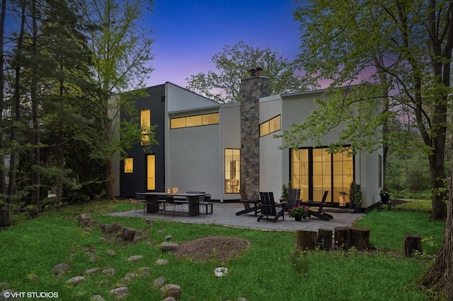 938 Old Green Bay Rd, Winnetka, IL 60093 - back of house at dusk featuring a chimney, a patio area, a lawn, and stucco siding