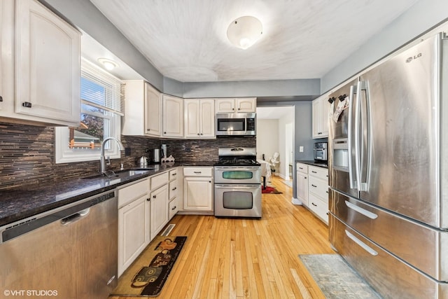 kitchen with stainless steel appliances, light wood finished floors, tasteful backsplash, a sink, and dark stone counters
