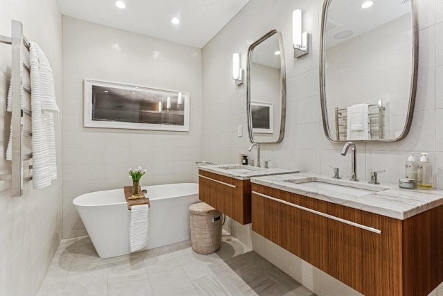 bathroom featuring tile walls, a soaking tub, two vanities, and recessed lighting