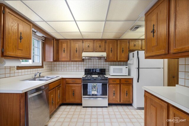 kitchen with white appliances, under cabinet range hood, brown cabinets, a paneled ceiling, and a sink