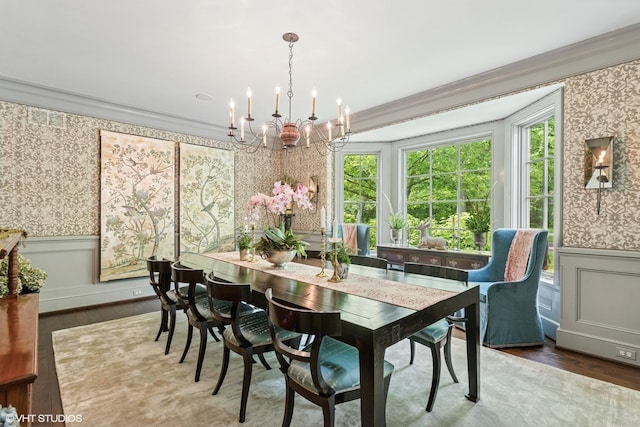 dining space featuring wood finished floors, wallpapered walls, a chandelier, crown molding, and a wainscoted wall