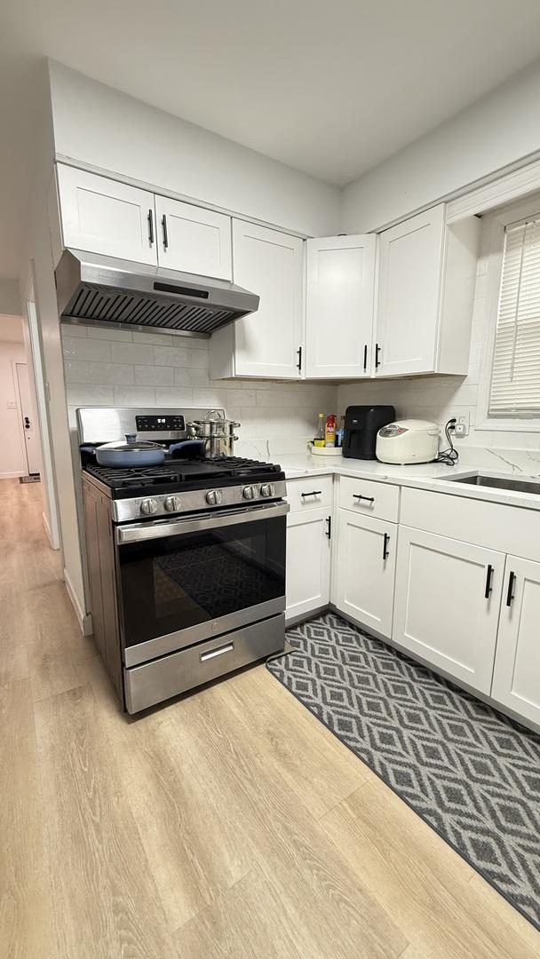 532 W 27th St, Chicago, IL 60616 - kitchen featuring stainless steel range with gas stovetop, under cabinet range hood, light wood-style flooring, light countertops, and white cabinetry