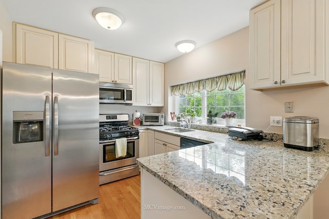 kitchen with stainless steel appliances, a peninsula, light wood-type flooring, cream cabinetry, and light stone counters
