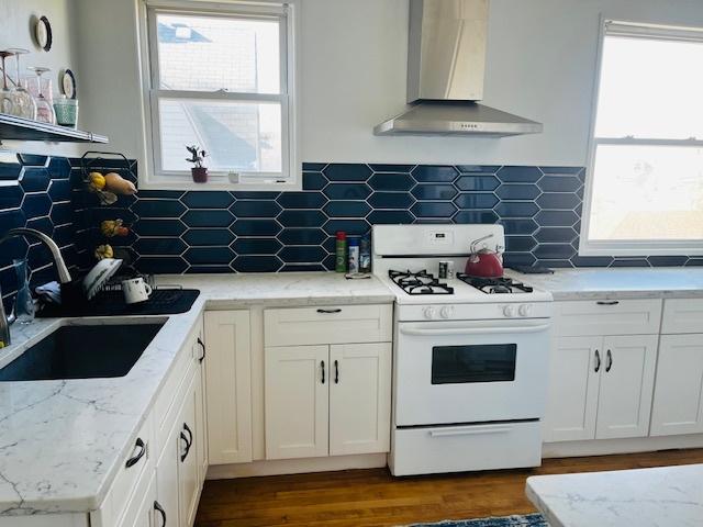 kitchen with white gas range oven, wall chimney exhaust hood, decorative backsplash, and white cabinetry