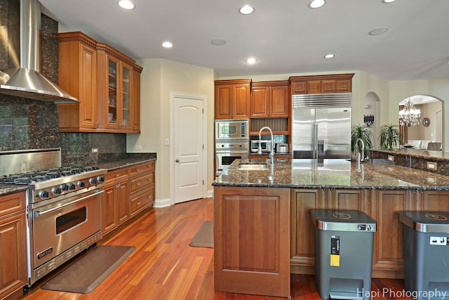 1400 Banbury Rd, Inverness, IL 60067 - kitchen featuring wall chimney range hood, built in appliances, arched walkways, brown cabinetry, and wood finished floors