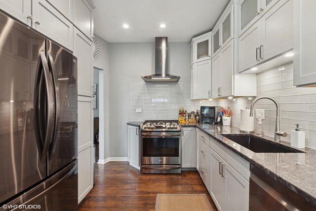 kitchen with appliances with stainless steel finishes, wall chimney range hood, dark stone counters, dark wood-style flooring, and tasteful backsplash