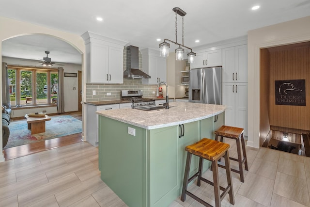 277 Mcmillan St, Grayslake, IL 60030 - kitchen featuring wall chimney exhaust hood, appliances with stainless steel finishes, a ceiling fan, a kitchen island with sink, and white cabinetry