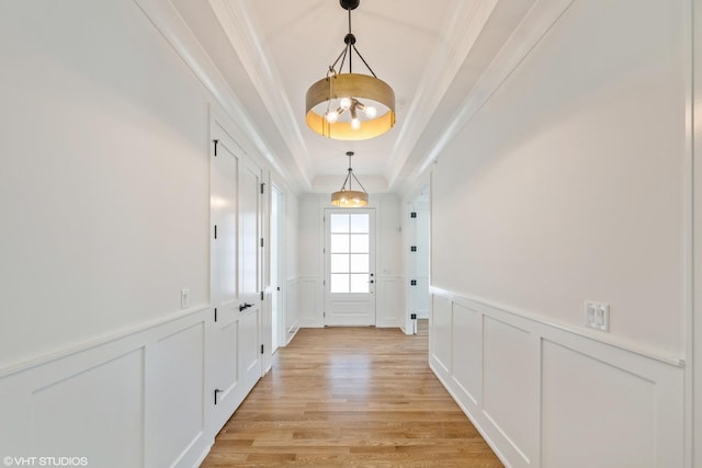 doorway to outside featuring a decorative wall, a raised ceiling, light wood finished floors, ornamental molding, and wainscoting