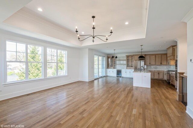 unfurnished living room with a raised ceiling, wine cooler, a chandelier, light wood finished floors, and recessed lighting