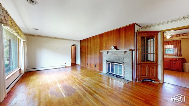 74 Wilmette Dr, Oswego, IL 60543 - unfurnished living room featuring baseboard heating, arched walkways, light wood-style floors, a stone fireplace, and wood walls