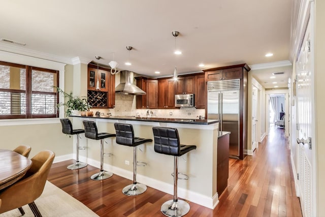 846 W Roscoe St Unit 3W, Chicago, IL 60657 - kitchen featuring stainless steel appliances, wall chimney exhaust hood, dark countertops, a breakfast bar area, and wood finished floors