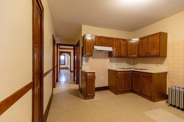 kitchen featuring under cabinet range hood, radiator, brown cabinets, light countertops, and tile walls