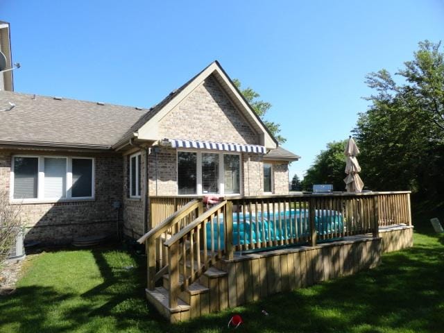 back of property featuring brick siding, a wooden deck, a yard, roof with shingles, and a covered pool