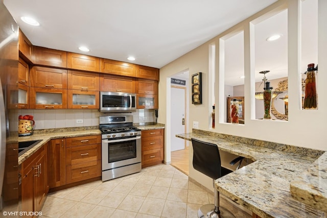 3300 N Carriageway Dr Unit 209, Arlington Heights, IL 60004 - kitchen featuring stainless steel appliances, brown cabinets, tasteful backsplash, glass insert cabinets, and light stone countertops