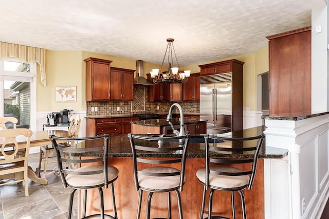 12499 Ayrshire Ln, Loves Park, IL 61111 - kitchen with built in fridge, wall chimney range hood, a chandelier, decorative backsplash, and a kitchen bar