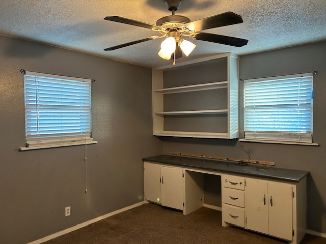 unfurnished office with baseboards, ceiling fan, dark carpet, built in desk, and a textured ceiling