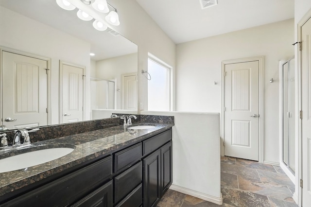 bathroom with stone tile floors, a shower stall, double vanity, and baseboards