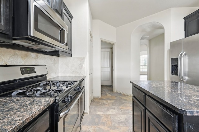 kitchen featuring appliances with stainless steel finishes, arched walkways, decorative backsplash, light stone finish flooring, and dark stone counters