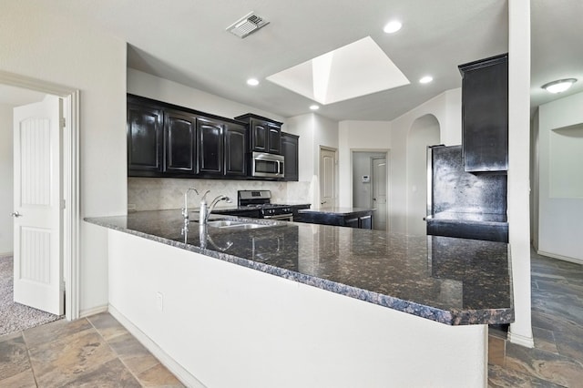 kitchen with stainless steel appliances, a sink, dark cabinetry, stone tile flooring, and a skylight