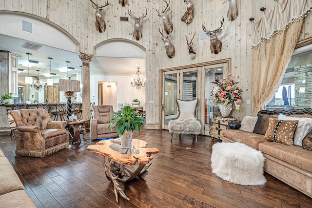 living room with a towering ceiling, decorative columns, dark wood-type flooring, wooden walls, and a notable chandelier