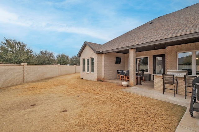 exterior space featuring a patio, a fenced backyard, brick siding, roof with shingles, and a bar