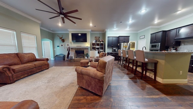 living room with arched walkways, a fireplace with raised hearth, ceiling fan, ornamental molding, and dark wood-type flooring