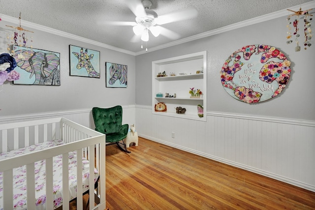 bedroom featuring ornamental molding, wainscoting, a textured ceiling, wood finished floors, and a nursery area