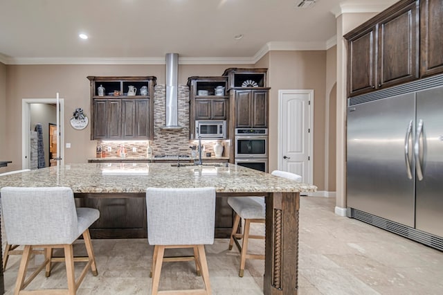 kitchen with built in appliances, light stone counters, a center island with sink, and wall chimney exhaust hood