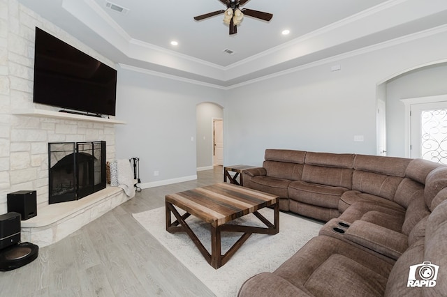 living room with ceiling fan, a raised ceiling, a stone fireplace, light hardwood / wood-style flooring, and crown molding