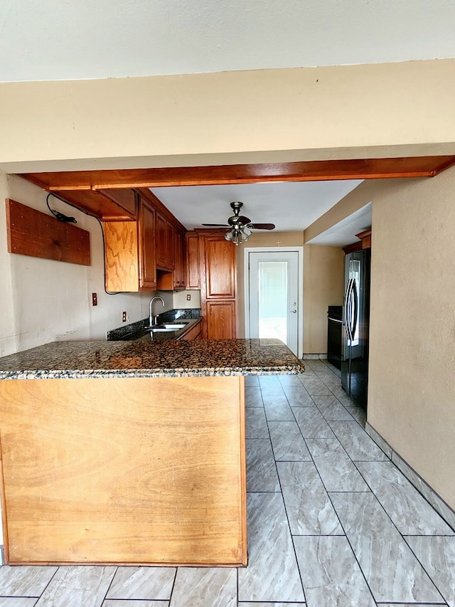 kitchen featuring sink, ceiling fan, dark stone countertops, kitchen peninsula, and stainless steel refrigerator