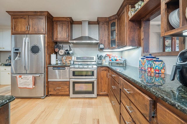 kitchen with wall chimney exhaust hood, light hardwood / wood-style floors, dark stone counters, and stainless steel appliances