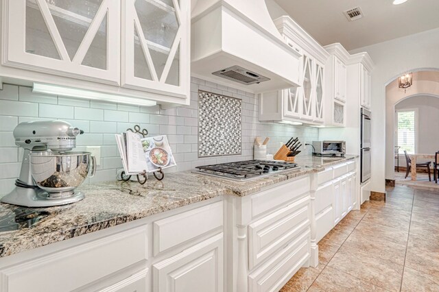 kitchen with stainless steel appliances, custom exhaust hood, arched walkways, and light stone counters