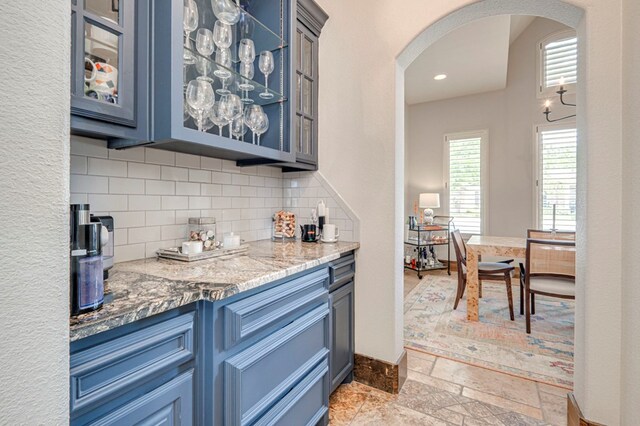 kitchen featuring arched walkways, blue cabinets, decorative backsplash, glass insert cabinets, and stone tile flooring