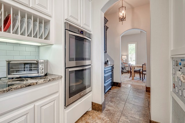 kitchen featuring stainless steel double oven, arched walkways, white cabinetry, tasteful backsplash, and light stone counters