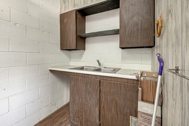 kitchen with a sink, concrete block wall, dark brown cabinetry, light countertops, and dark wood finished floors