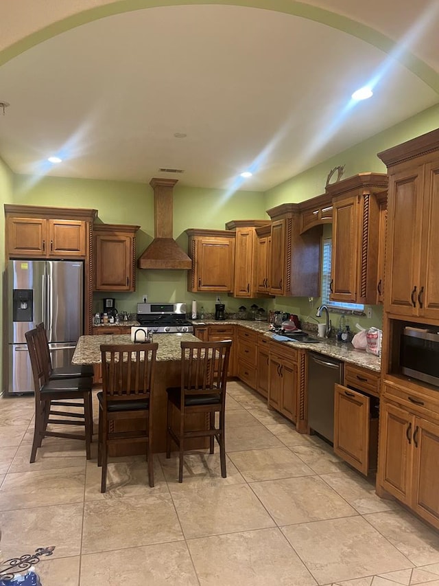 kitchen with a breakfast bar, a sink, appliances with stainless steel finishes, wall chimney range hood, and light stone countertops