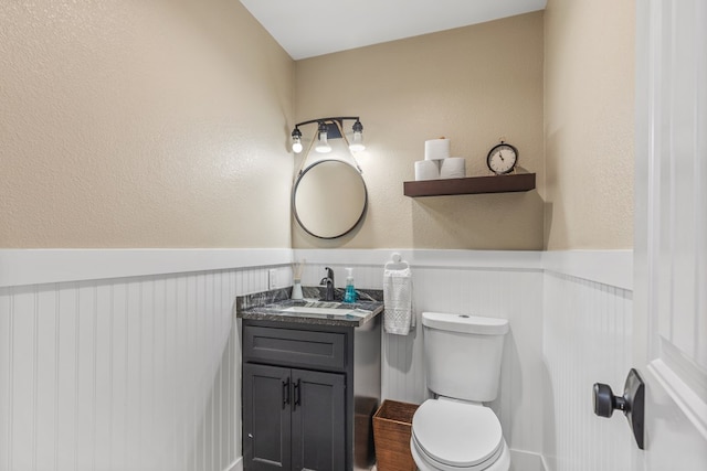 half bath with a wainscoted wall, a textured wall, toilet, and vanity