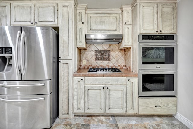 2406 Stutz Pl, Midland, TX 79705 - kitchen featuring stainless steel appliances, under cabinet range hood, cream cabinetry, decorative backsplash, and tile counters