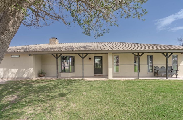 back of house featuring stucco siding, a chimney, a patio area, a lawn, and metal roof
