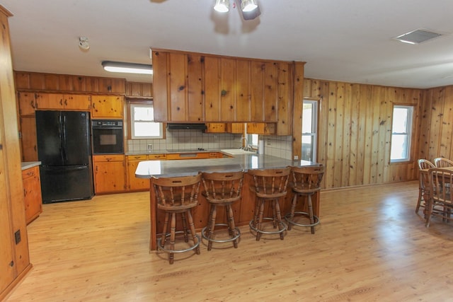 kitchen featuring wooden walls, light hardwood / wood-style flooring, a breakfast bar area, decorative backsplash, and black appliances