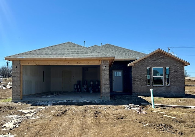 unfinished property featuring a patio area, brick siding, roof with shingles, and an attached garage